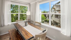 A dining room with a wooden table, white chairs, a vase, and a view of a residential area through large windows. - Spacious & Modern 3 Bedroom in Madison Valley (Seattle)