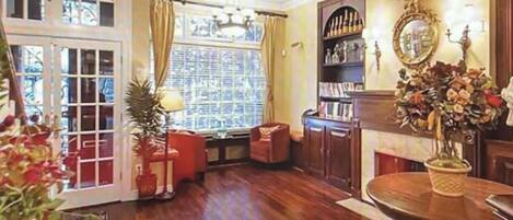 Una habitación con piso de madera, chimenea, estantería y ventana con cortinas.