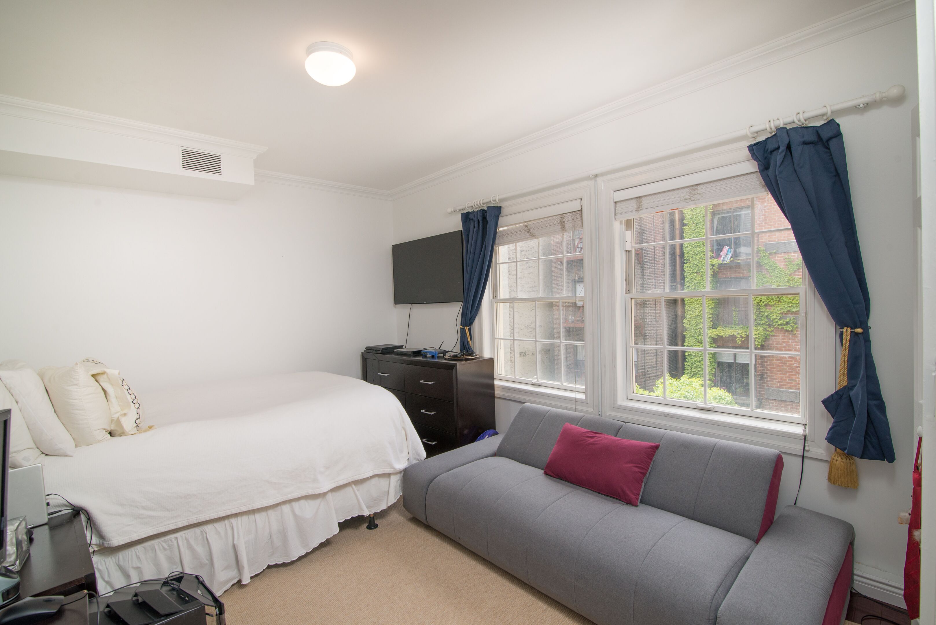 Un dormitorio con cama, un sofá gris, televisor en la pared y ventana con cortinas.