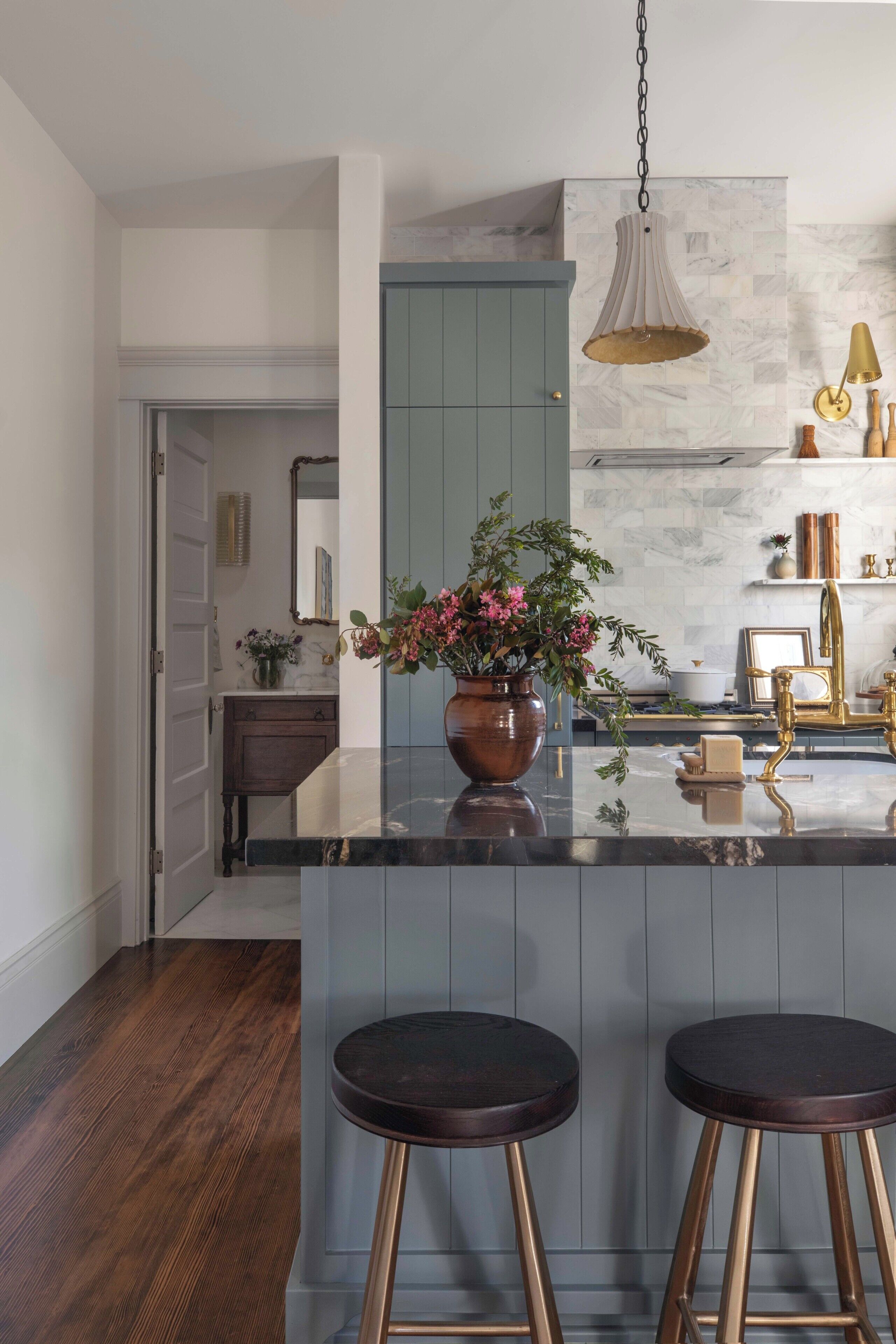 A modern kitchen with a marble countertop, two stools, a hanging light fixture, and a vase with flowers.