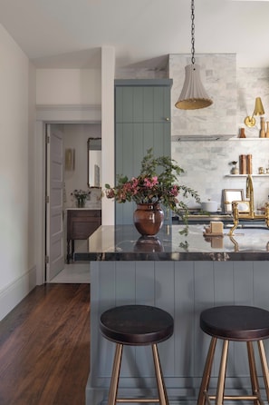 A modern kitchen with a marble countertop, two stools, a hanging light fixture, and a vase with flowers.
