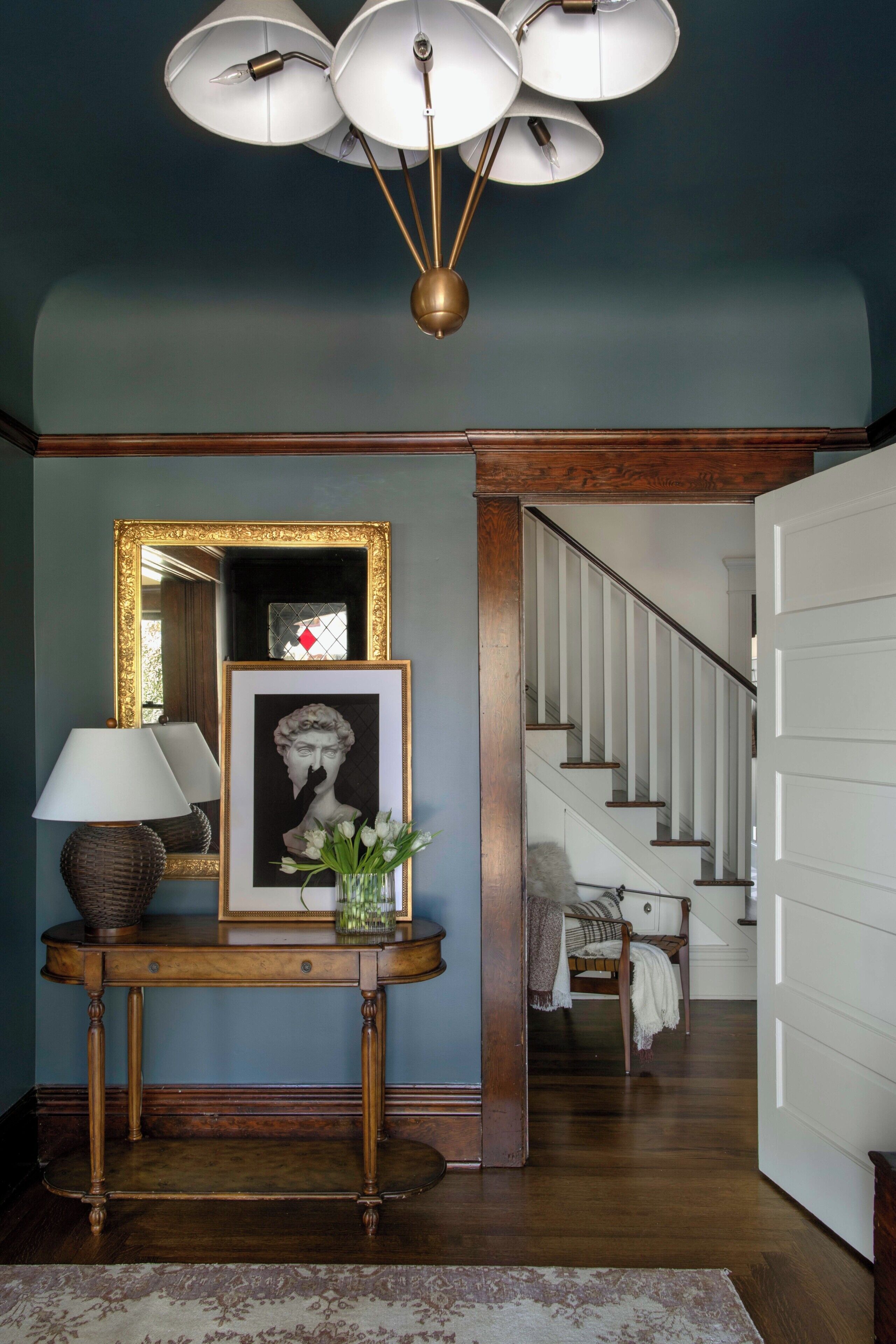A hallway with a console table, a framed artwork, a lamp, and a staircase leading to another room.