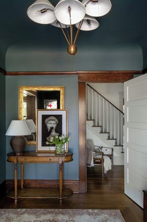A hallway with a console table, a framed artwork, a lamp, and a staircase leading to another room. - The Craft House Inn (Santa Barbara)