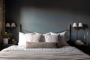 A bedroom with a bed, pillows, a nightstand, a lamp, and a vase with flowers. - The Craft House Inn (Santa Barbara)