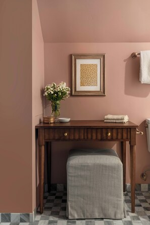 A bathroom with a wooden vanity, a framed floral artwork, a vase with white flowers, and a towel rack with a white towel. - The Craft House Inn (Santa Barbara)