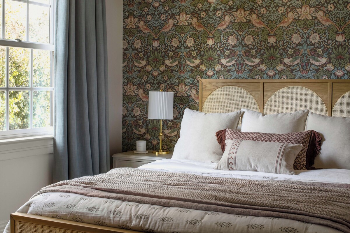 Quarto com cama, mesa de cabeceira, abajur e janela com cortinas.