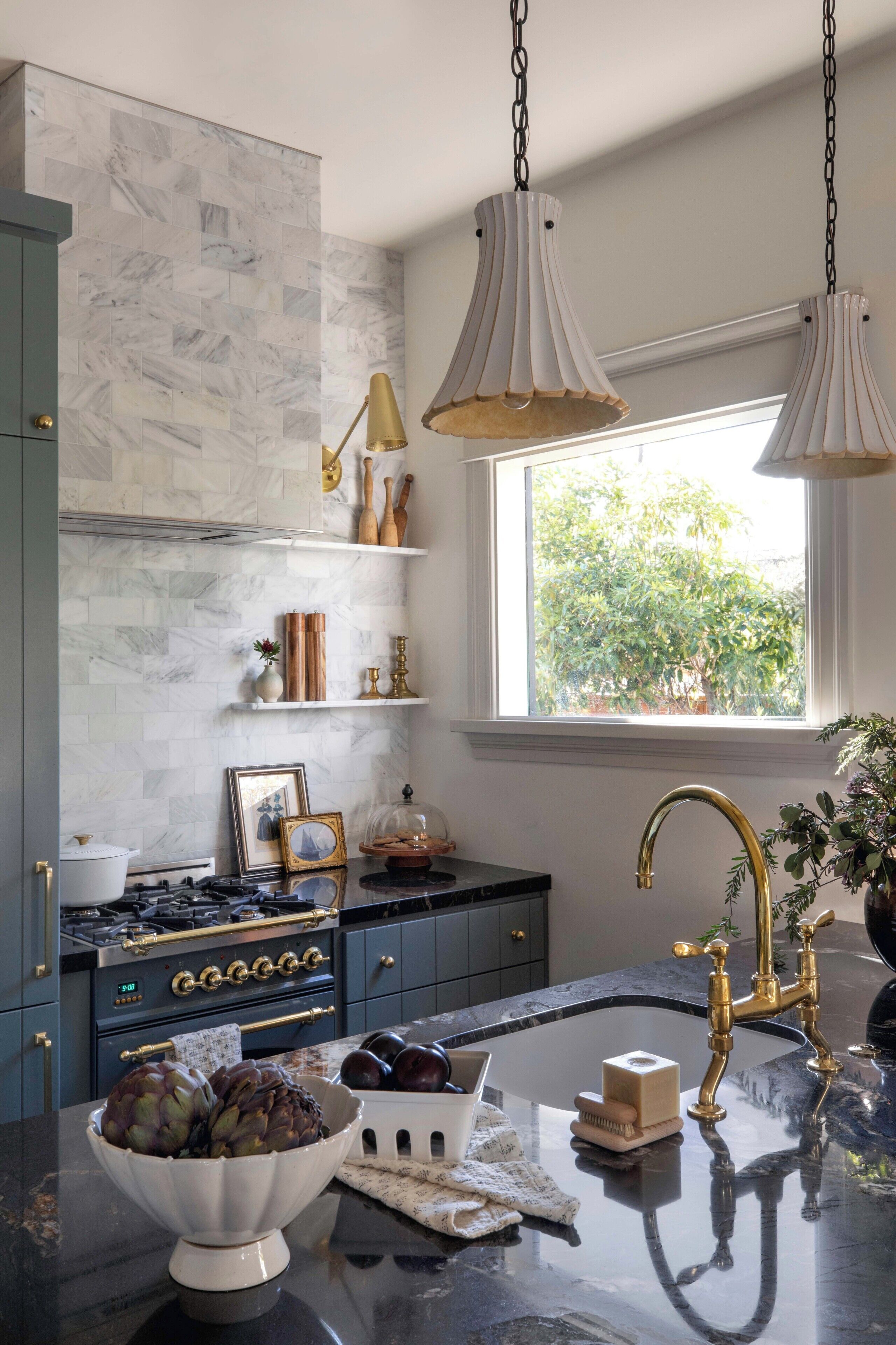 A modern kitchen with marble backsplash, a gas stove, a sink with gold fixtures, and a window with a view of greenery.