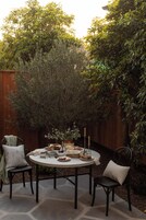 An outdoor patio with a round table set for a meal, surrounded by chairs, with a wooden fence and foliage in the background.