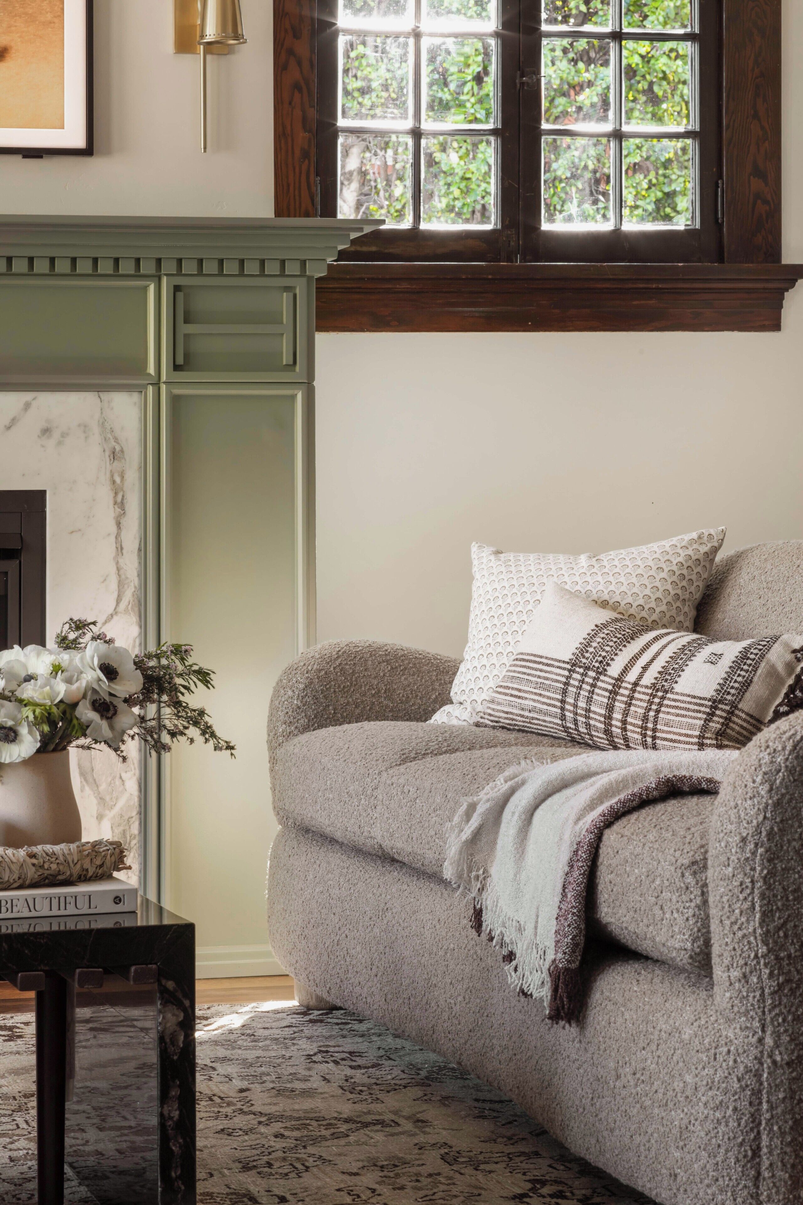 A cozy living room with a sofa, a vase with flowers, and a window with a view of greenery.