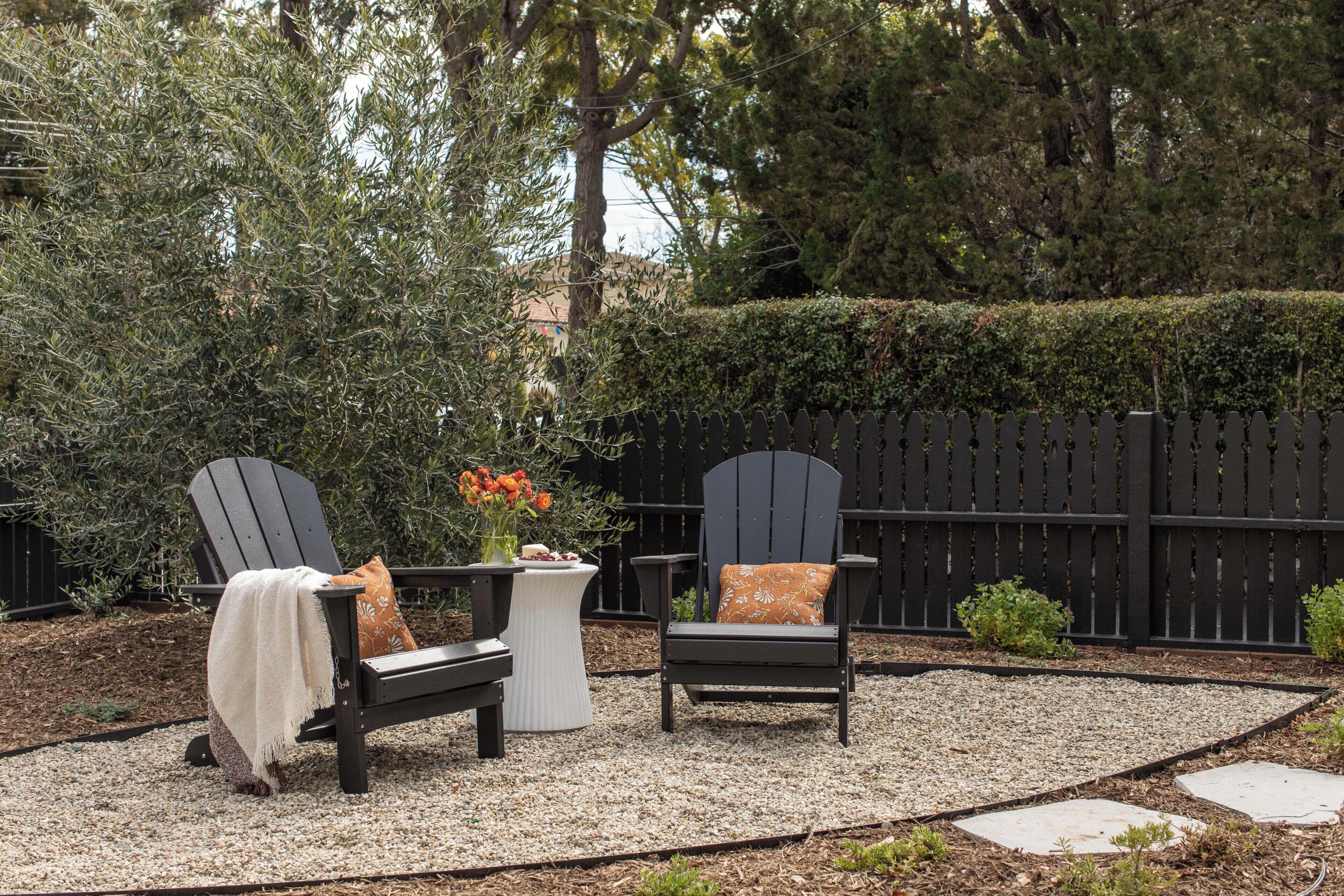 Dos sillas Adirondack con cojines, una mesa redonda pequeña con un florero y una zona de patio con grava.
