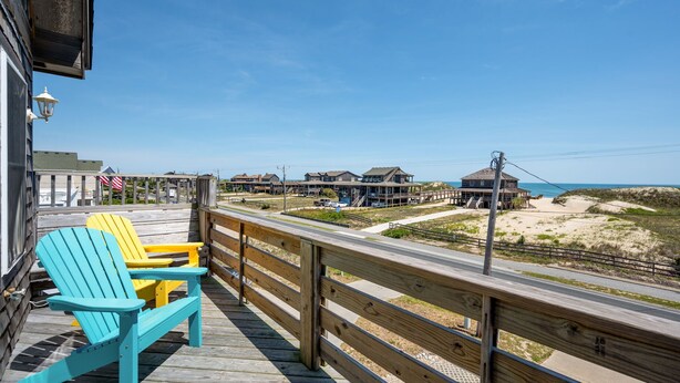 A wooden deck with two Adirondack chairs, overlooking a beach with buildings and a clear sky. - Footloose & Fab-Semi-oceanfront, pool, hot tub, rec room, pool table, elevator! (Nags Head)