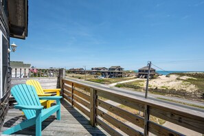 A wooden deck with two Adirondack chairs, overlooking a beach with buildings and a clear sky.
