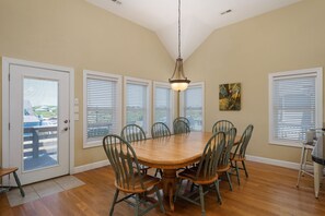 A dining room with a wooden table and chairs, large windows with blinds, and a hanging light fixture. - Footloose & Fab-Semi-oceanfront, pool, hot tub, rec room, pool table, elevator! (Nags Head)