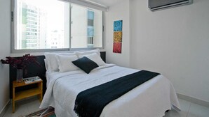 A bedroom with a large window, a bed with white bedding and a dark throw, a nightstand with a lamp and books, and a wall-mounted air conditioner.