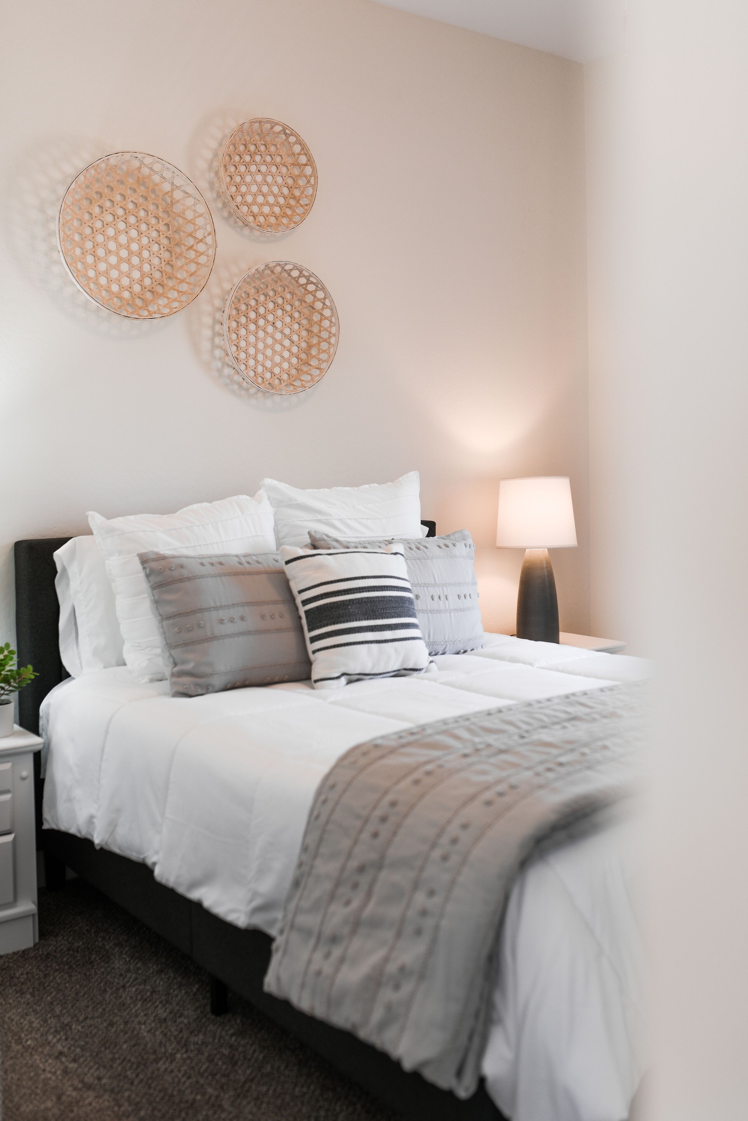 A neatly made bed with white bedding and multiple pillows, a bedside lamp, and three wall-mounted woven baskets.