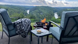 Ein Tisch mit Feuer, zwei Gläsern Rotwein und einer Schüssel mit Snacks, mit Blick auf einen Fluss und bewaldete Hügel.