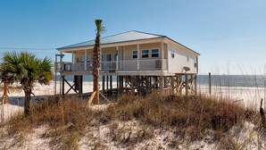 A beach house with a balcony, located on stilts over the sand, with the ocean in the background.