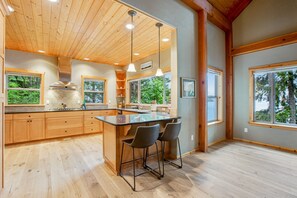 Cocina amplia con armarios de madera, una isla central con taburetes y grandes ventanales con vistas al verdor.