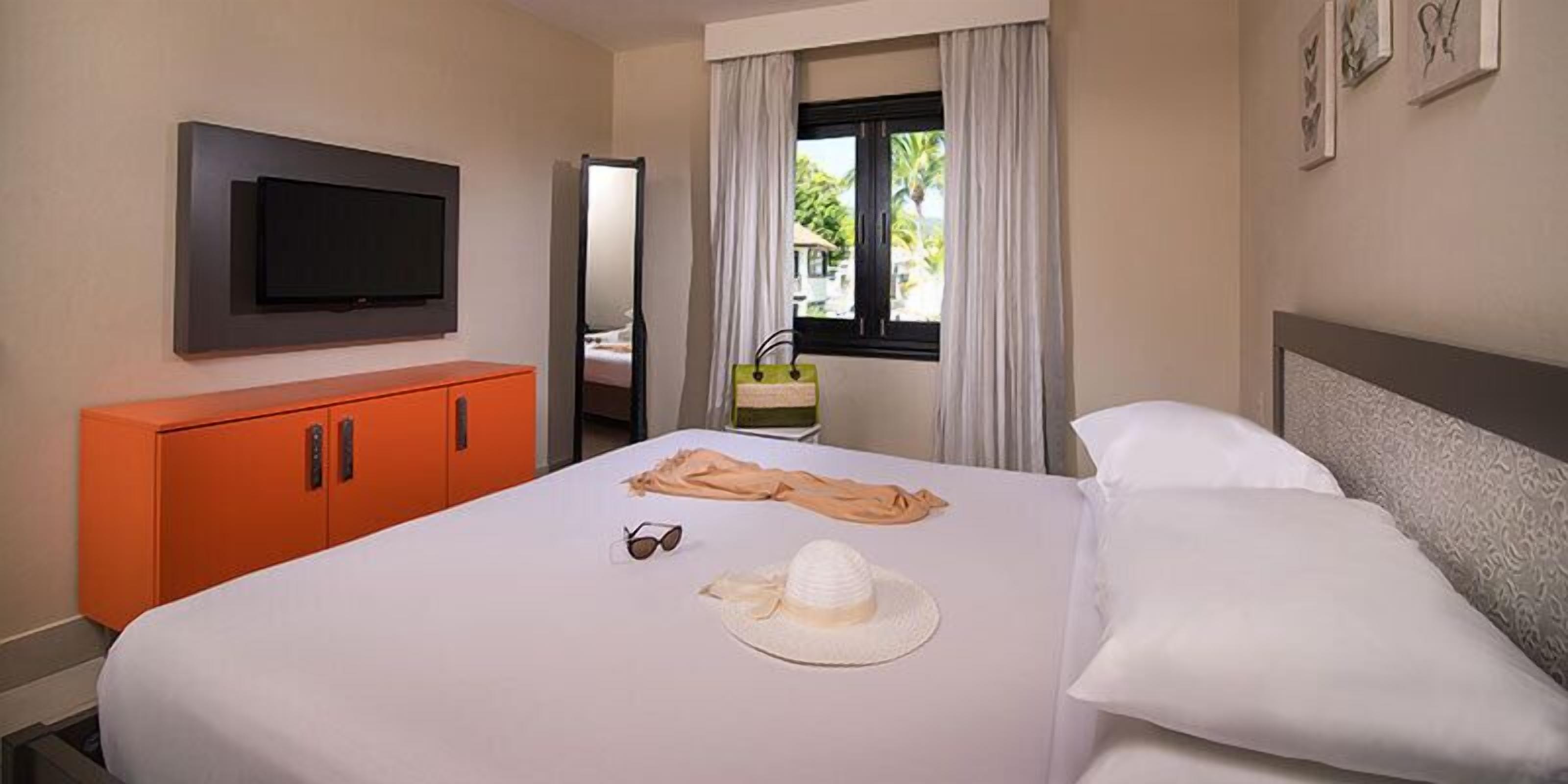 Una habitación de hotel con una cama, un televisor montado en la pared, un armario naranja y una ventana con cortinas.