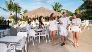 An outdoor dining area with white tables and chairs, a thatched-roof structure, and palm trees. - Cofresi Puerto Plata Dominican Republic  (Puerto Plata)