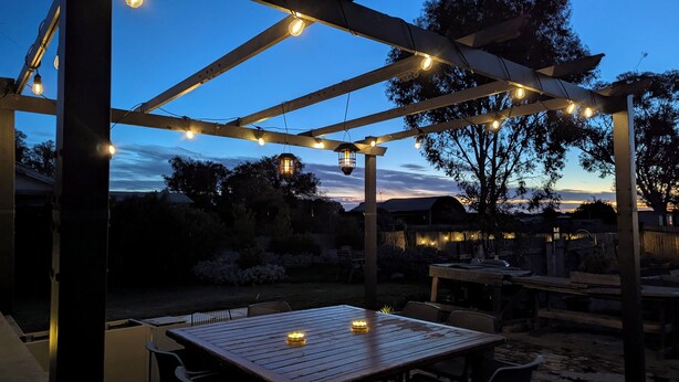Een buitenpatio met een houten pergola, lichtjes en een tafel met kaarsen.