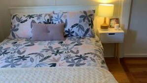 Een bed met een bloemenprint deken en een witte sprei. Er staat een nachtkastje met een lamp en een klein fotolijstje.