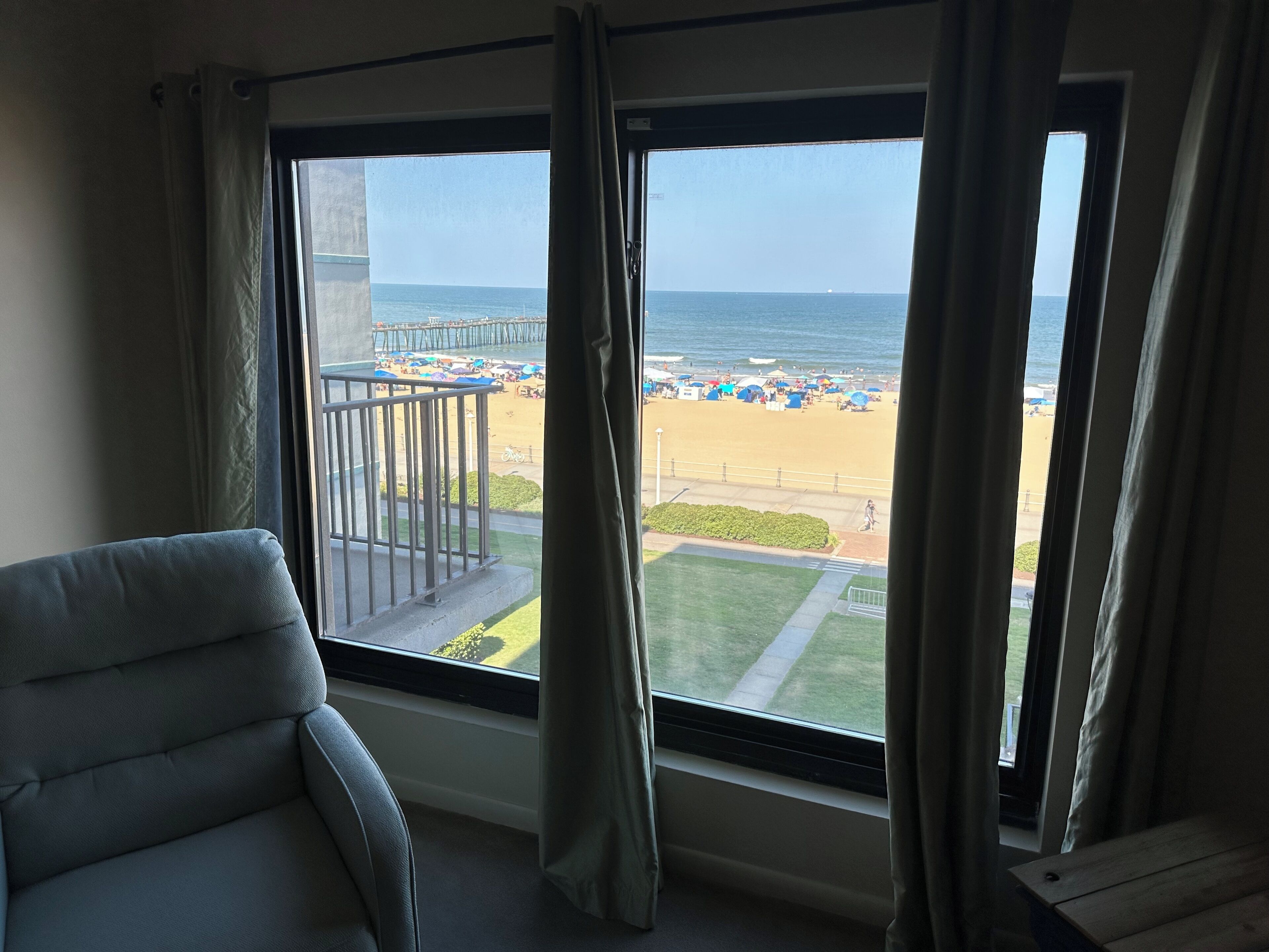 A room with a large window overlooking a beach, a balcony with a railing, and a chair.