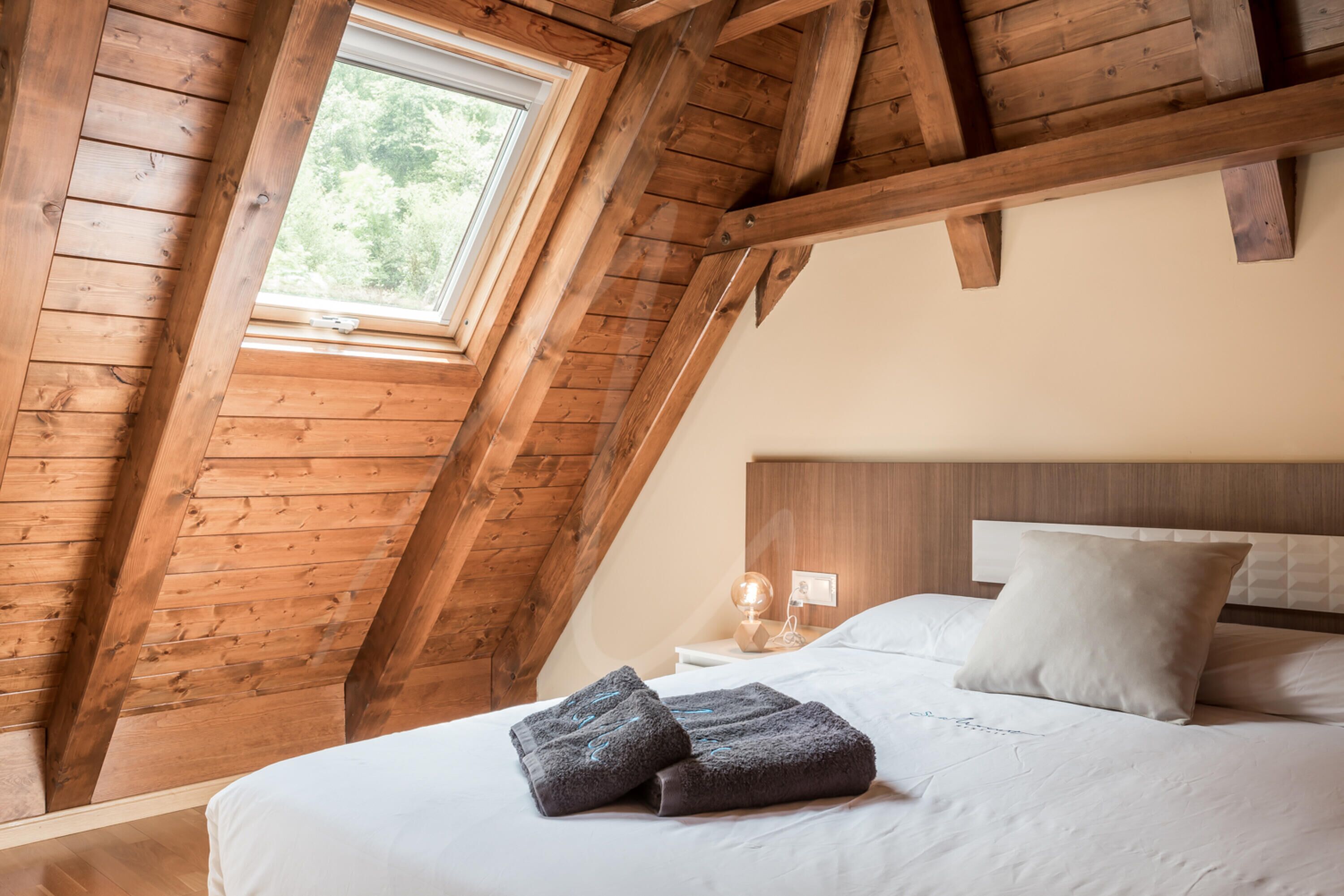 Ein Schlafzimmer mit schrägen Wänden, einem Bett mit weißer Bettwäsche, einem Nachttisch mit Lampe und einem Fenster mit Blick auf Grünflächen.