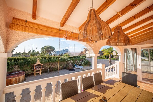 Un área de comedor al aire libre techada con mobiliario de mimbre y lámparas colgantes, con vistas a un jardín ajardinado y una carretera.