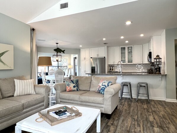 A spacious living room with a beige sofa, a white coffee table, and a kitchen area with white cabinets and a wooden countertop.
