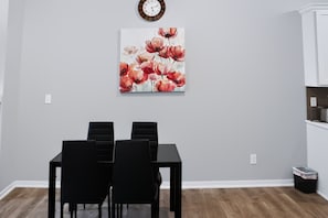 A dining area with a table and chairs, a clock on the wall, and a painting of flowers.