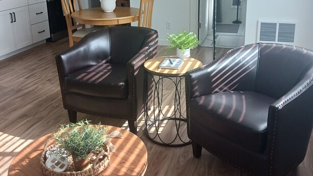 A modern living room with a wooden coffee table, two leather armchairs, a dining table with chairs, and a kitchen in the background.