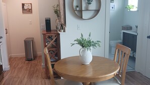 A dining area with a round wooden table, four chairs, and a white vase with green plants. There is a wooden cabinet with a shelf above the table, and a framed picture on the wall.