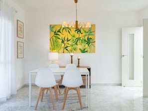 A dining area with a white table, four white chairs, a yellow painting with green leaves, and a hanging light fixture.
