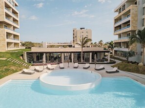 Una piscina amb gandules, una zona coberta amb ombrelles i un edifici amb balcons al fons.
