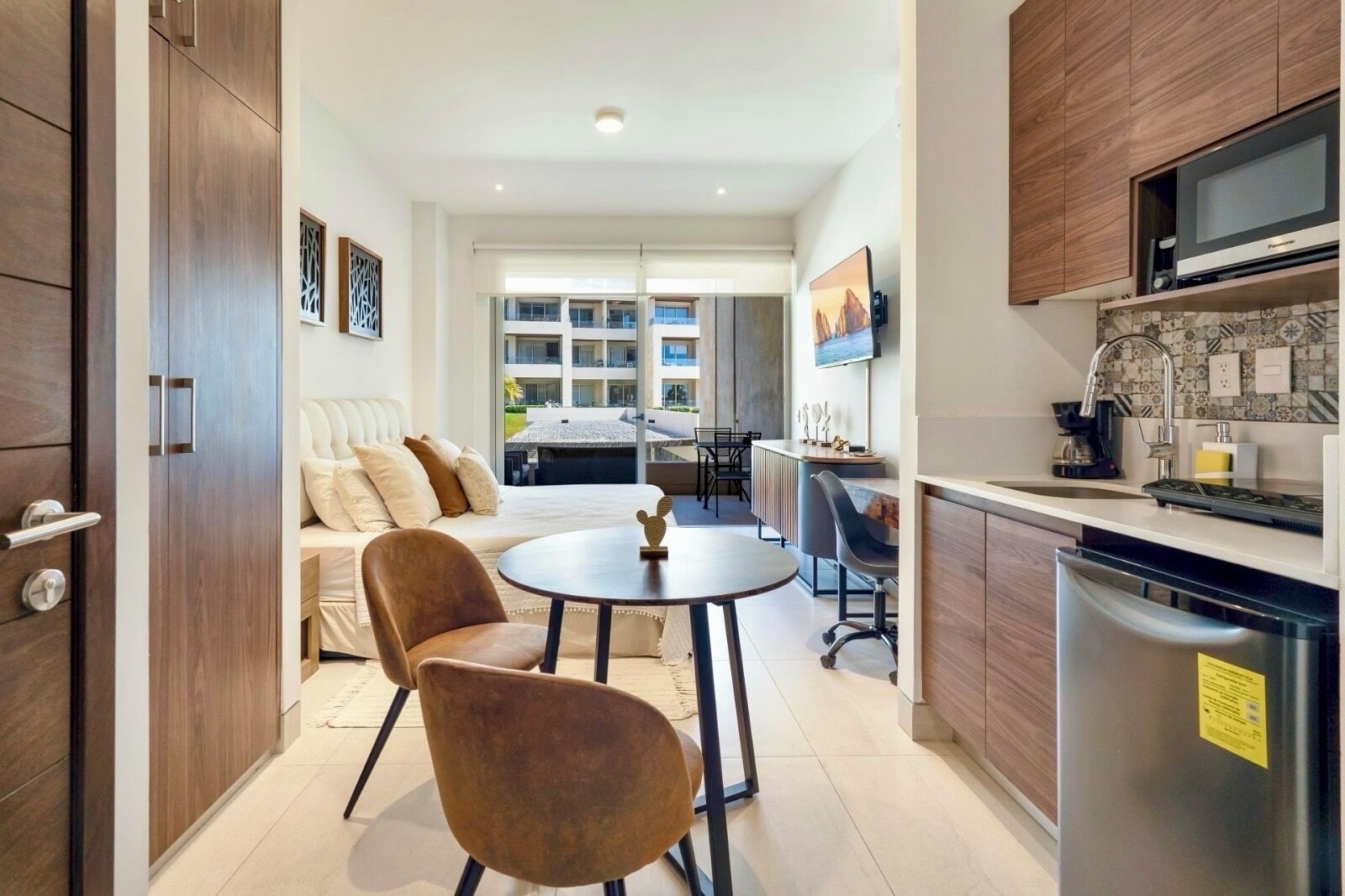 A modern hotel room with a sofa, two chairs, a small table, a kitchen area with a refrigerator, microwave, and sink.