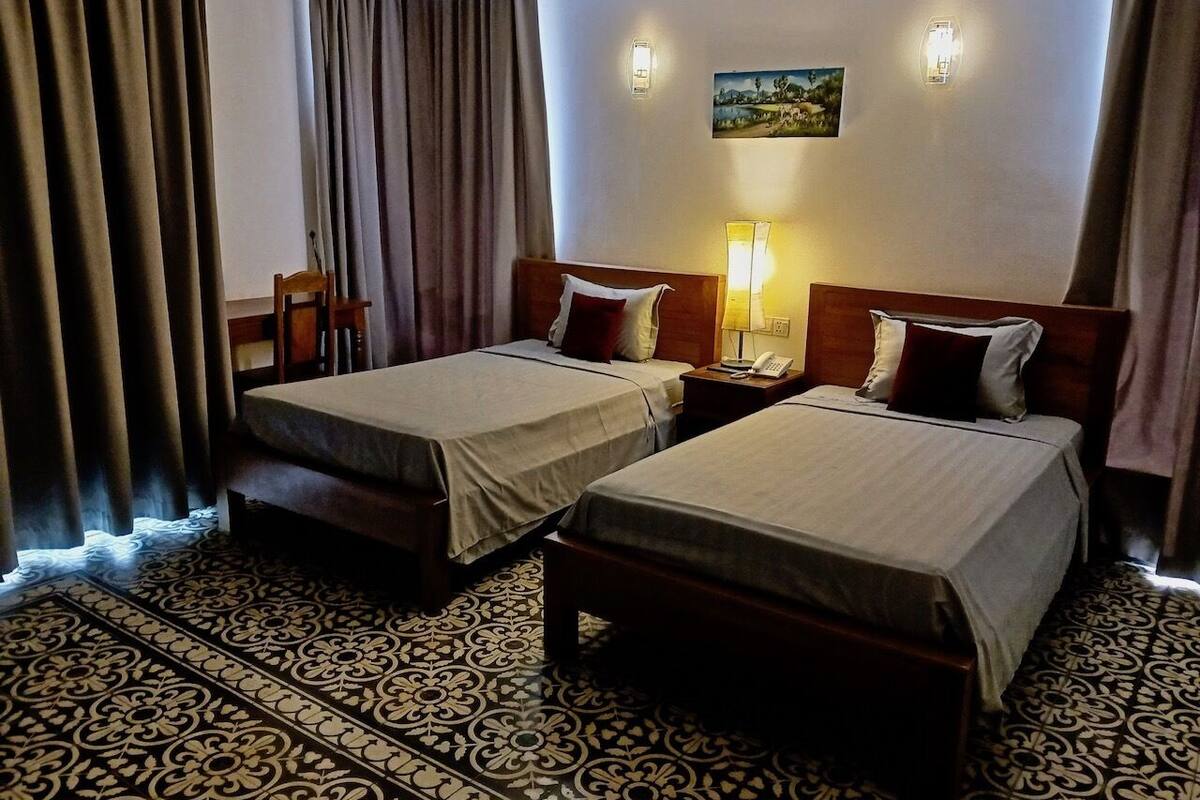 Une chambre d’hôtel avec deux lits simples, une moquette à motifs, un tableau au mur et une table de chevet avec une lampe.