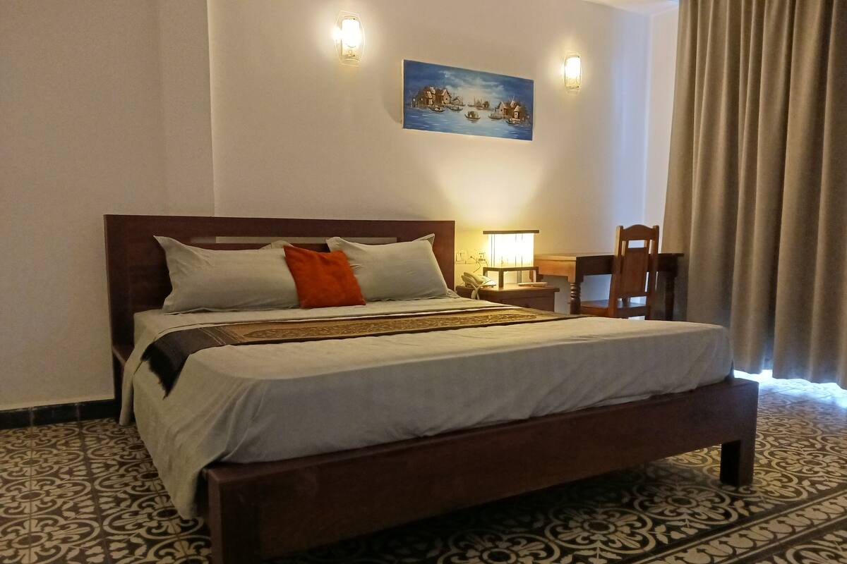 Un lit bien fait, avec une tête de lit en bois, une table de chevet avec une lampe, une chaise et un tableau accroché au mur.