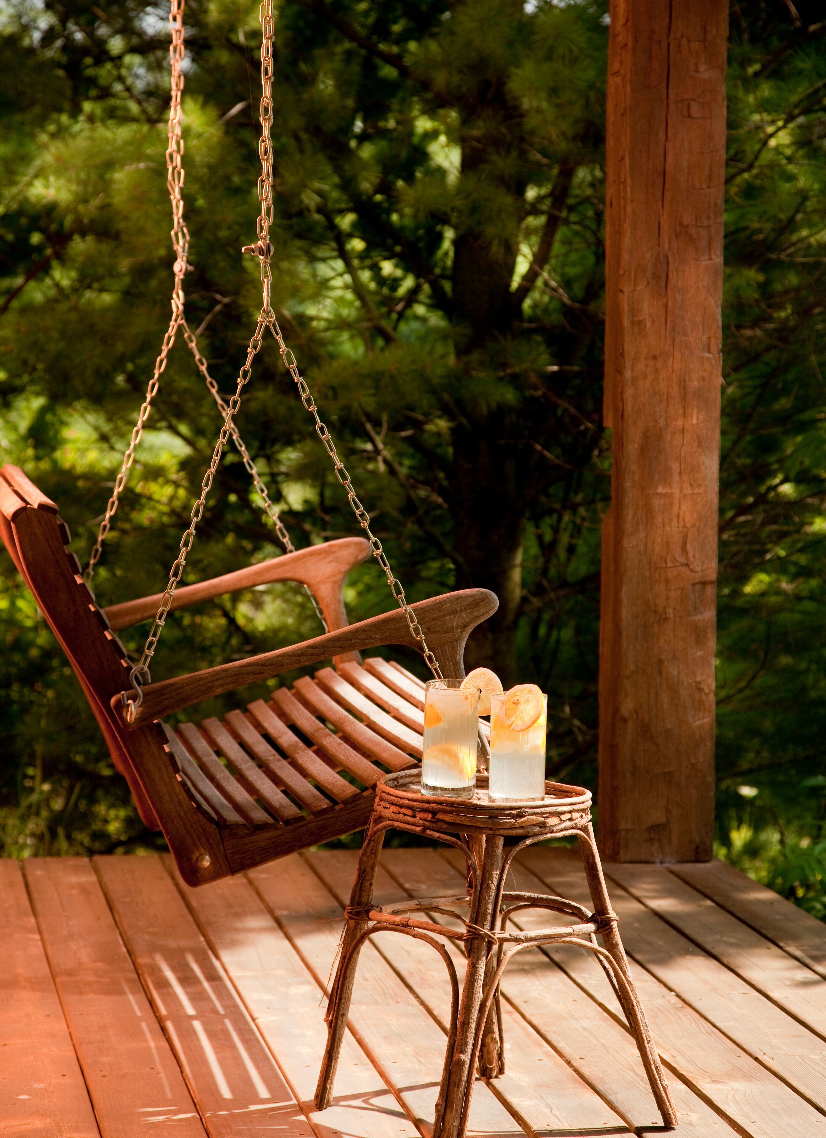 A wooden swing chair suspended by chains, a small table with two glasses of a clear beverage, and a wooden bench.