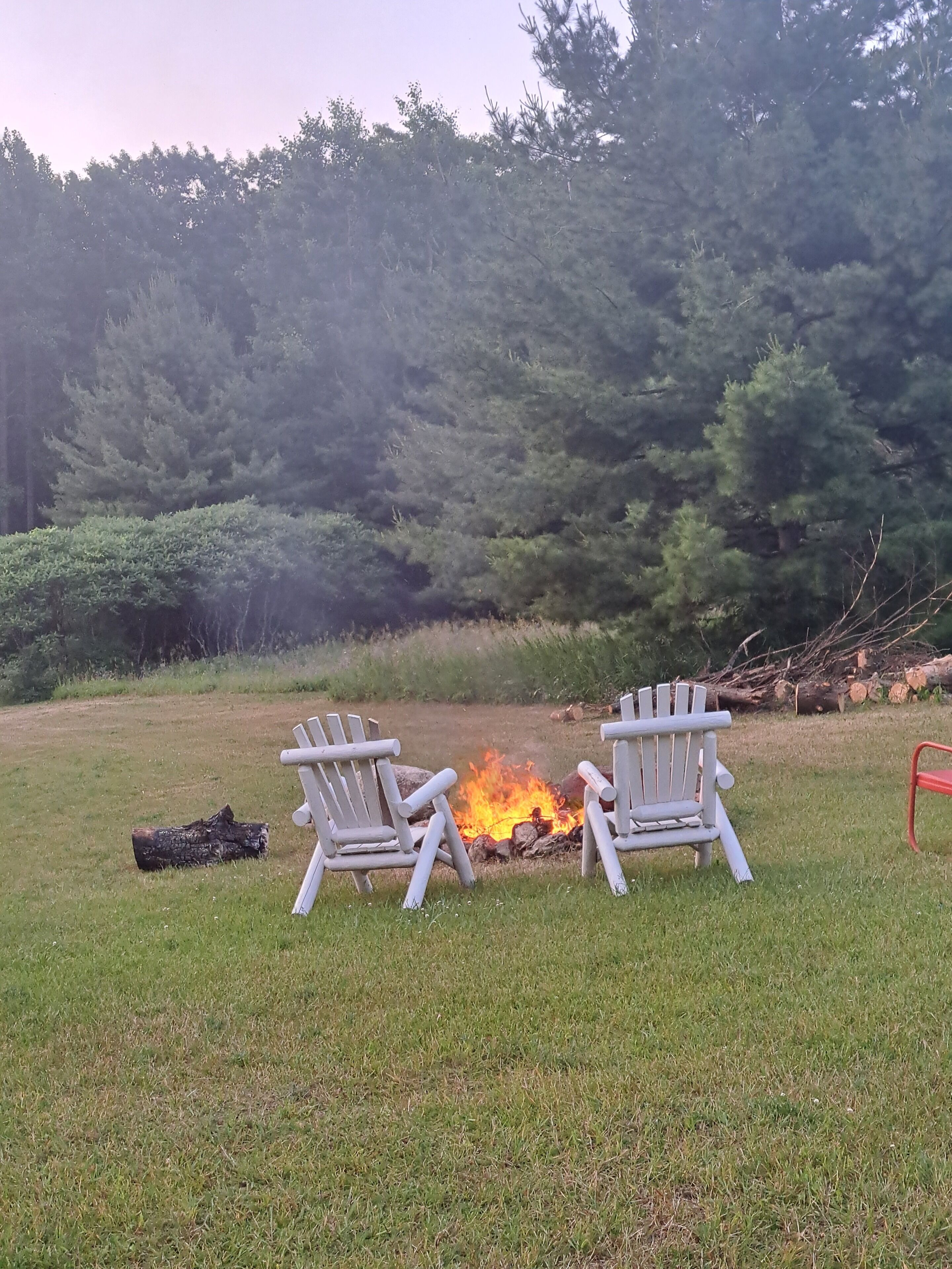 Two Adirondack chairs facing a campfire in a grassy field with trees in the background.