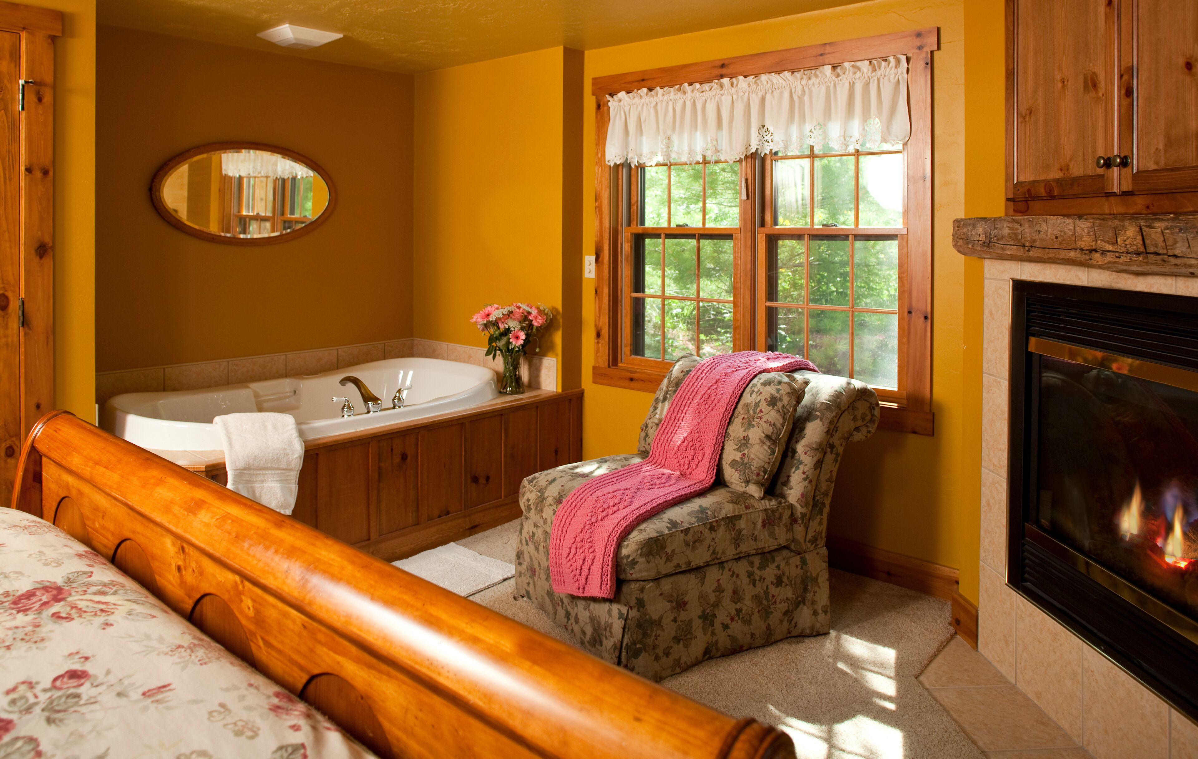 A bedroom with a wooden bed, a bathroom with a bathtub, a fireplace, and a window with a view of trees.