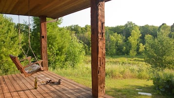 A wooden deck with a swing chair, a bottle, and a glass on the table, overlooking a forested area.