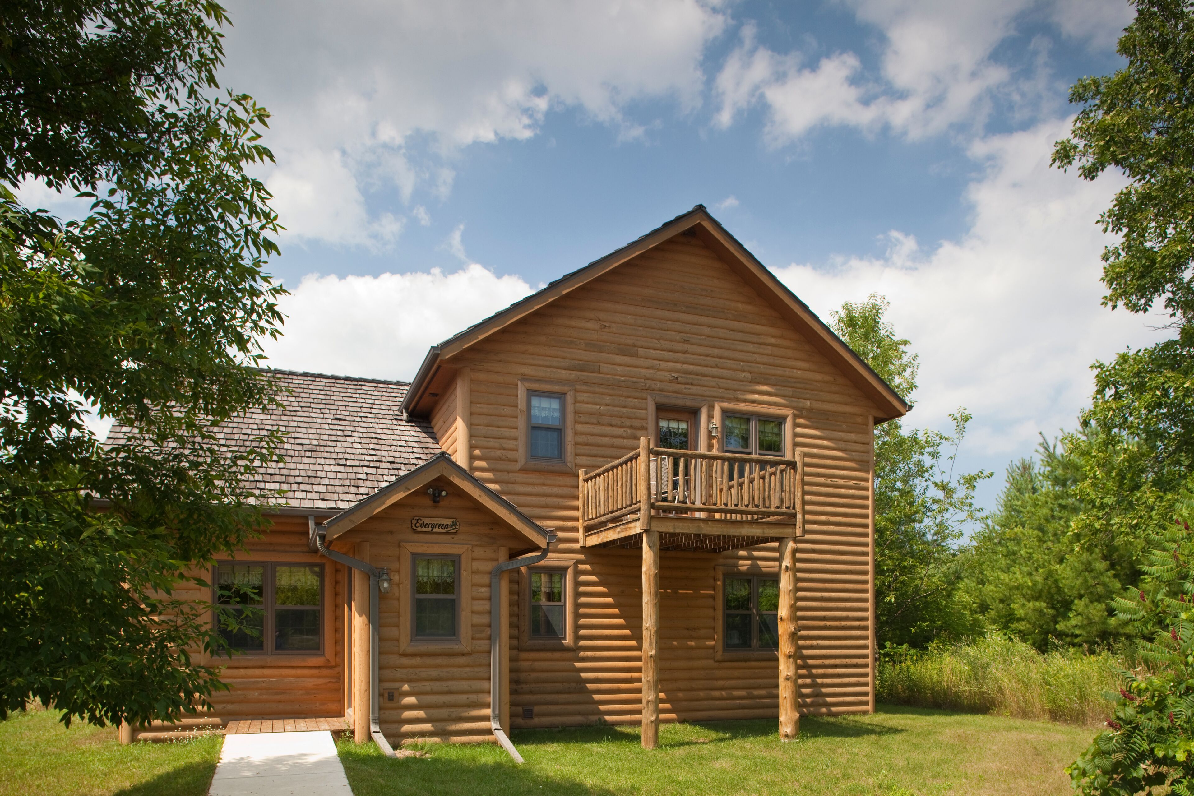 A wooden log cabin with a balcony, a front porch, and a small attached structure.