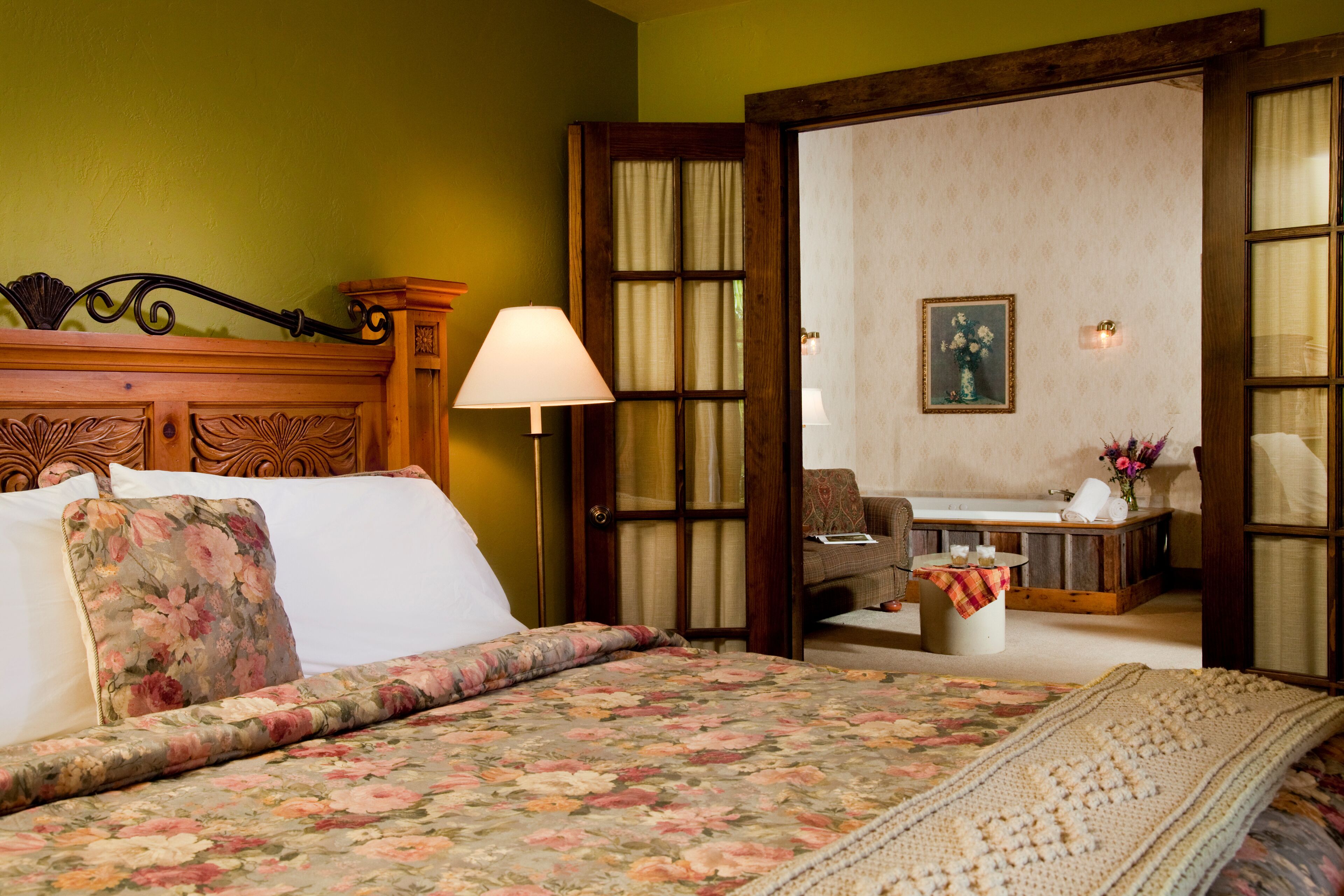 A bedroom with a floral bedspread, a wooden headboard, a bedside lamp, and a view of a dining area through an open door.