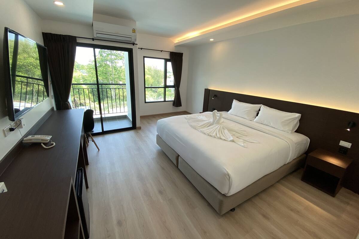 A modern hotel room with a large bed, a desk with a telephone, and a balcony with a view of greenery.