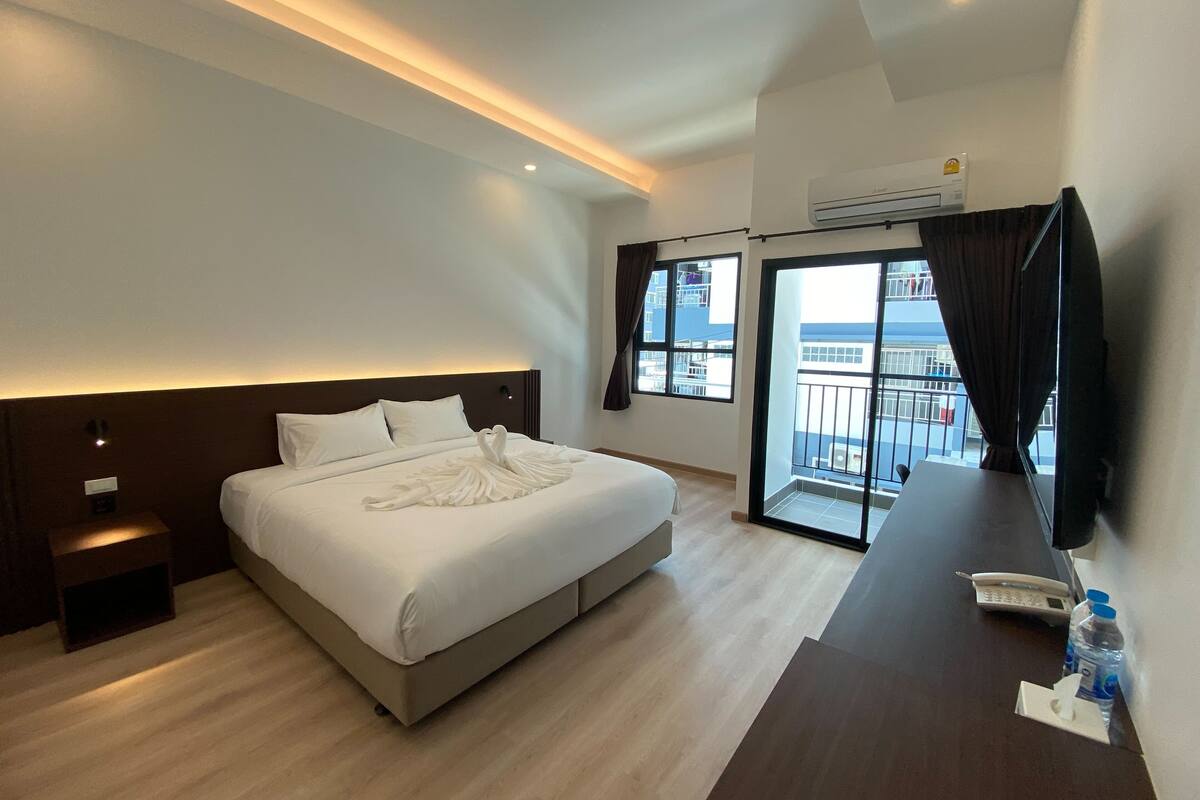 A modern hotel room with a large bed, a desk with a telephone and water bottle, a balcony with a view, and an air conditioning unit.