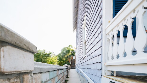 Ein schmaler Betonweg, der von einer Backsteinmauer und einem weißen Geländer mit blauen Fensterläden gesäumt wird.