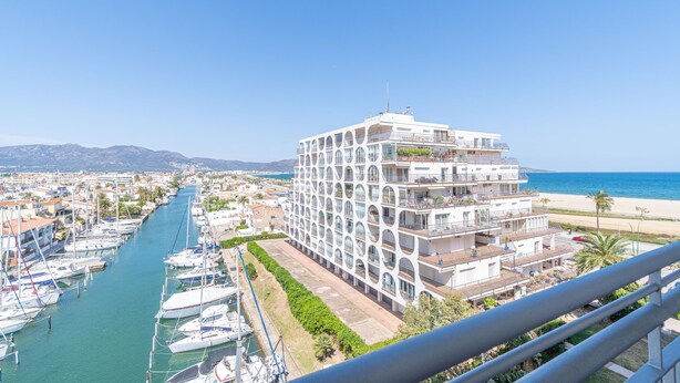 Een jachthaven met boten, een modern wit gebouw met balkons, en een strand op de achtergrond.