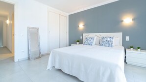 Een slaapkamer met een groot bed, witte muren en een grijze accentmuur. Er staan twee nachtkastjes met lampen en een klein plantje op elk.