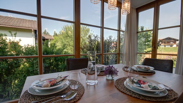 Una mesa de comedor dispuesta con platos, cubiertos y cristalería, con vistas a zonas verdes y un edificio a través de amplios ventanales.